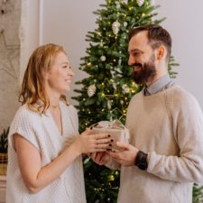 Woman giving man a holiday gift by Christmas tree.