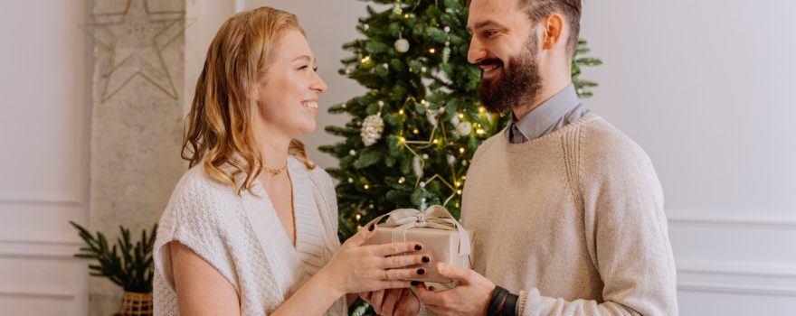 Woman giving man a holiday gift by Christmas tree.