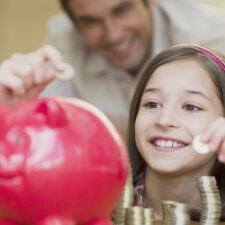 Father and daughter putting coins in a piggie bank, which can be a way of teaching kids financial literacy.
