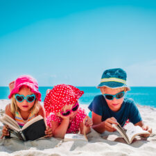 Three young children reading books on a beach. Books for your kids' summer reading list include stories about animals, school and more.