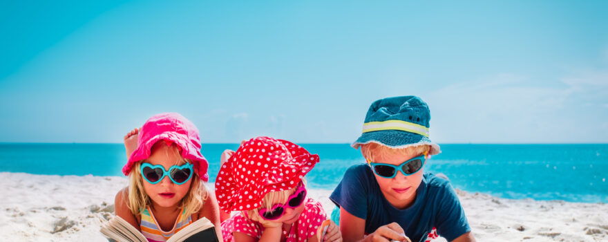 Three young children reading books on a beach. Books for your kids' summer reading list include stories about animals, school and more.