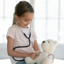 child using a stethoscope on a teddy bear; now is a great time to make appointments for back-to-school checkups
