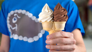 person wearing a Fudgie the Whale shirt while holding a cone with both vanilla and chocolate ice cream, part of Carvel's National Soft Serve Day deals.