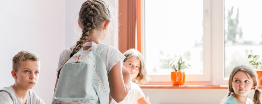 Four kids in a classroom near a window with sun coming through. One is standing and looks like she's starting a new school or class.