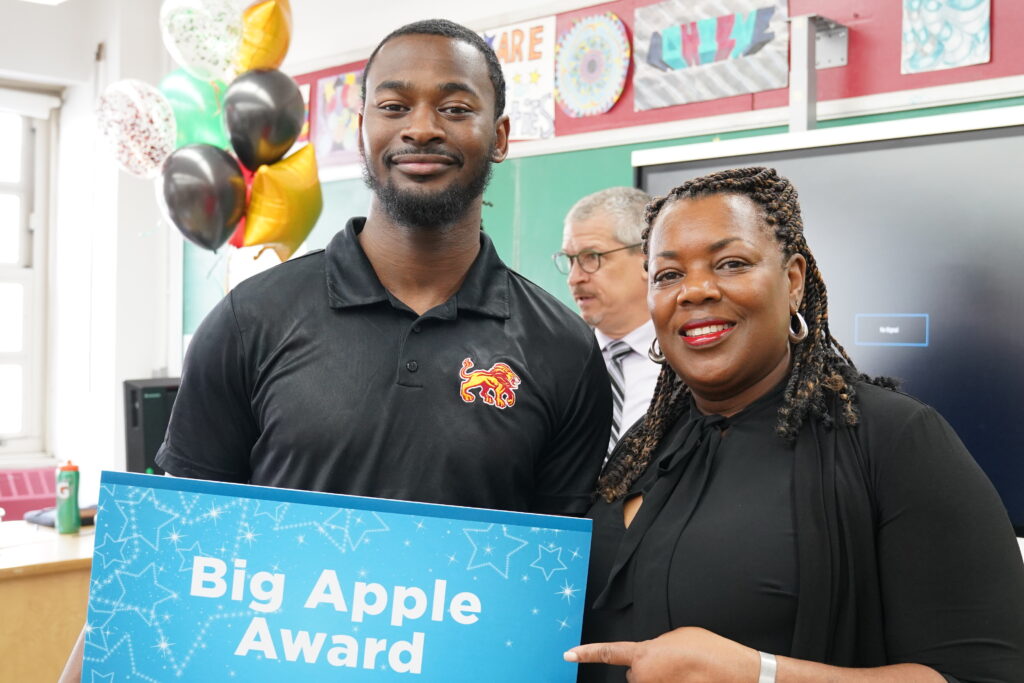two people holding a Big Apple Awards sign in a classroom 